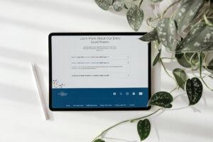 A tablet with a stylus beside it displays a David Weekley Homes webpage about entry-level homes, placed on a white surface near green leafy plants. The screen shows text and dropdown menus with placeholder content.