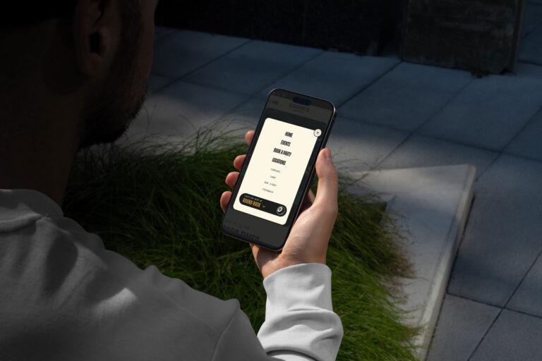 A person holding a smartphone outdoors, looking at a screen displaying a list of options with a highlighted button that says NEXT STEP—perhaps planning their visit to Eddie's Tavern. Sunlight and greenery fill the background. | Terrain