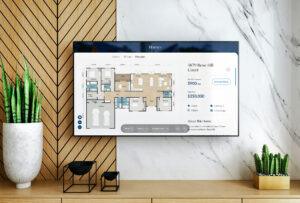A TV Kiosk mounted on a marble wall displays an Imagination Homes floor plan and listing details. Next to the TV, potted plants and decorative items sit on a wooden shelf with a diagonal pattern. | Terrain