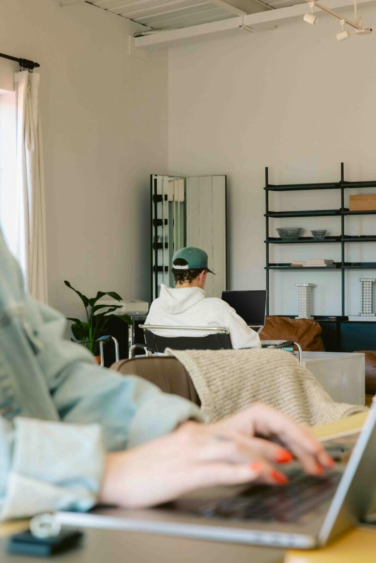 Person in foreground with red nail polish types on a laptop, possibly working on graphic design, while another person in a cap and hoodie sits at a desk in a modern, cozy room filled with shelves and plants. | Terrain