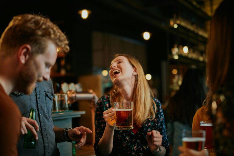 A woman holding a beer mug laughs joyfully while socializing with friends at a lively, dimly lit bar—capturing the perfect scene for any vibrant brewery website design. Other patrons hold drinks and chat, adding to the cheerful atmosphere. | Terrain
