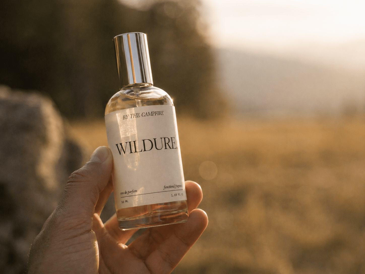 A hand holds a glass bottle of perfume labeled WILDURE BY THE CAMPFIRE against a blurred outdoor background with golden sunlight. | Terrain