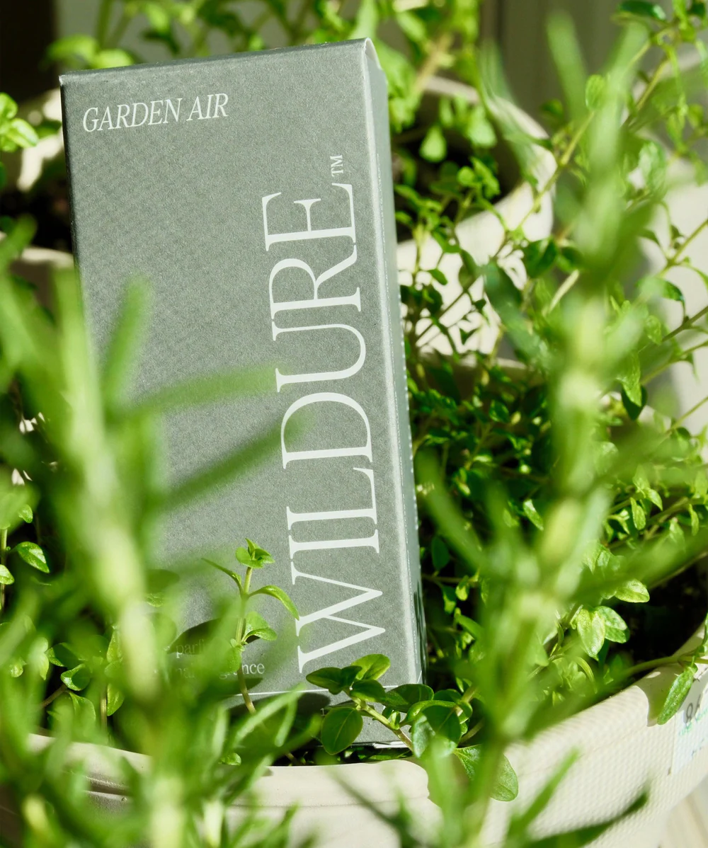A gray box labeled WILDURE Garden Air stands upright among lush green potted plants, partially obscured by leaves in the foreground. | Terrain