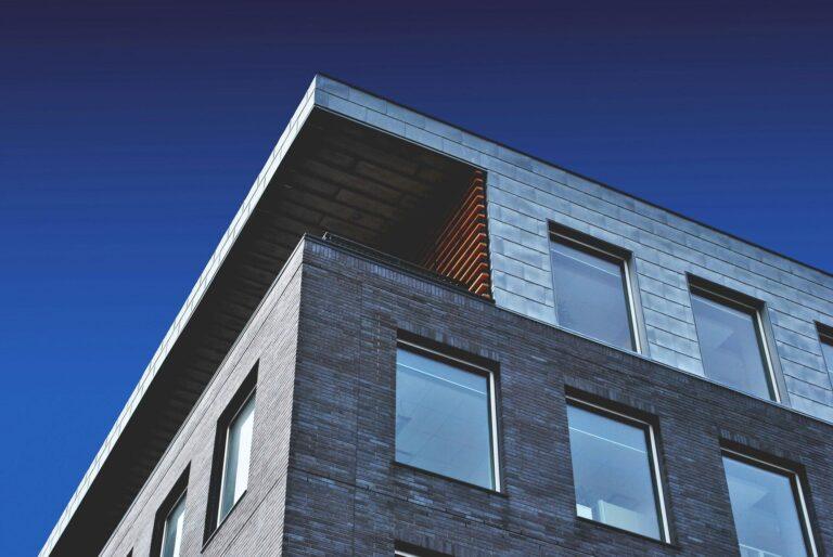 A modern building with large windows and a gray brick facade, set against a clear deep blue sky—perfect for showcasing on a real estate developer website. Its angular, minimalist design features sleek metal cladding on the upper section. | Terrain