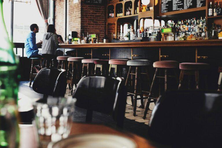 A cozy bar with empty stools at the counter, where two people chat beneath shelves of bottled drinks and glassware. Tables fill the foreground—an inviting scene perfect for showcasing on your restaurant website in 2026. | Terrain