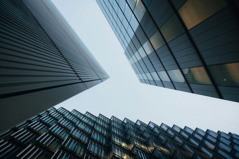 A low-angle view of three modern office buildings with glass and steel exteriors, converging towards the sky—an ideal visual for a 2026 commercial real estate firm website, framed against a pale, overcast background. | Terrain