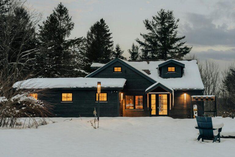 A dark wooden house with glowing windows sits in a snowy landscape at dusk, surrounded by tall trees. An empty blue Adirondack chair is in the foreground, while soft lights evoke the charm often found on the best builder websites. | Terrain
