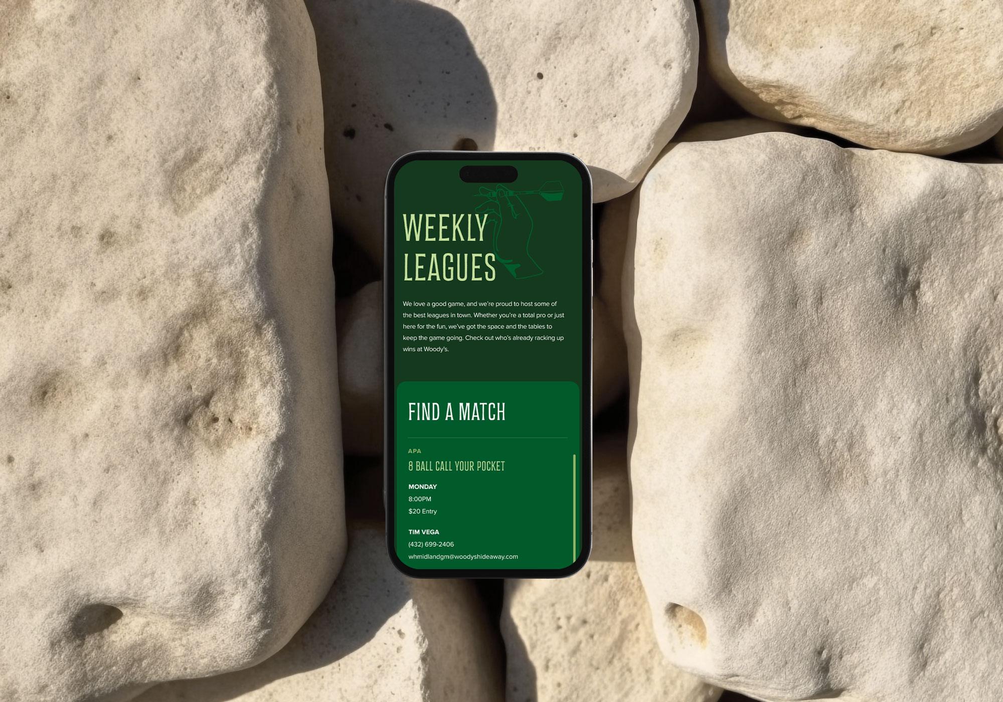 A smartphone with a green screen displaying Woody's Hideaway Weekly Leagues pool tournament info rests on large, smooth white rocks in sunlight. The screen features event details and contact information in white and yellow text. | Terrain