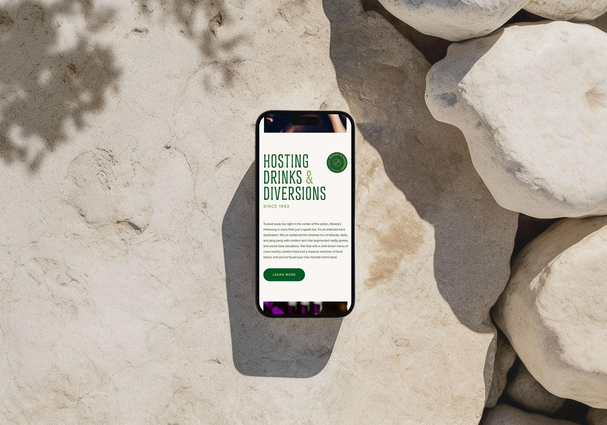A smartphone rests on light-colored rocks, displaying a website titled Hosting Drinks & Diversions with a green button labeled Learn More. Shadows of leaves cover the rocks, hinting at the relaxed vibe of Woody's Hideaway. | Terrain